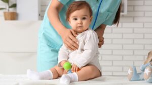 A baby in a white outfit sits on an exam table holding a toy, while a healthcare professional in scrubs gently supports them from behind. Urgent Care Omaha